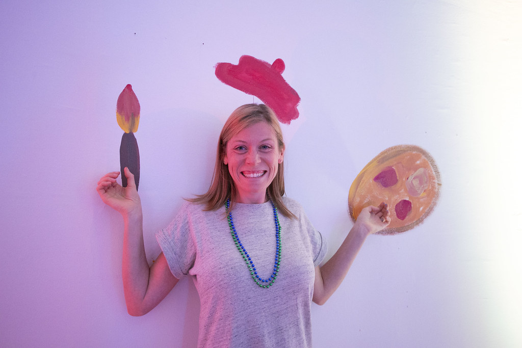 Photo of a woman wearing a gray t-shirt smiling and standing in front of a while wall painted to look like she is holding a paintbrush and artist palette.