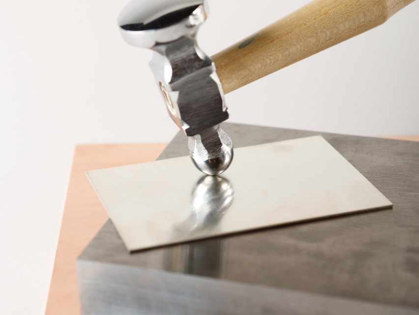 Close-up photo of a hammer hitting a small sheet of silver metal on top of a larger metal block.