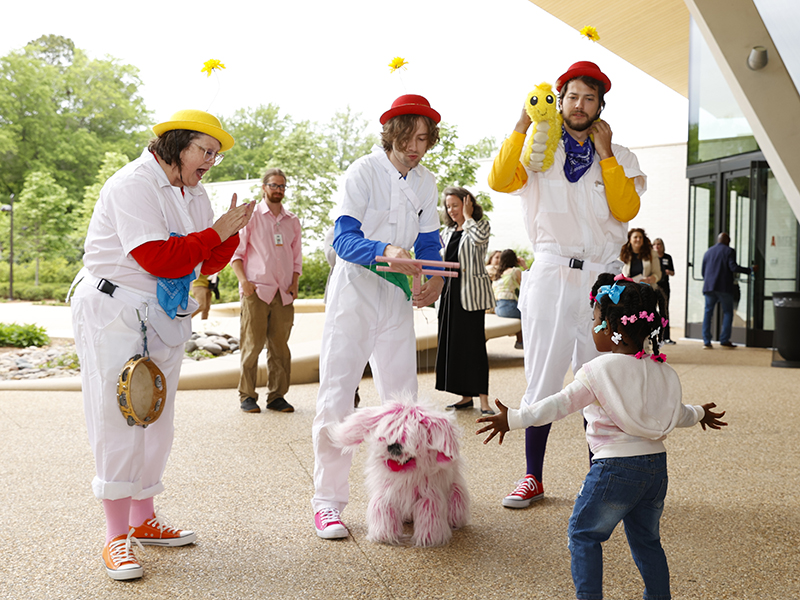 Photo of three puppeteers in white jumpsuits holding a pink dog puppet and yellow worm puppet while interacting with a small young child.