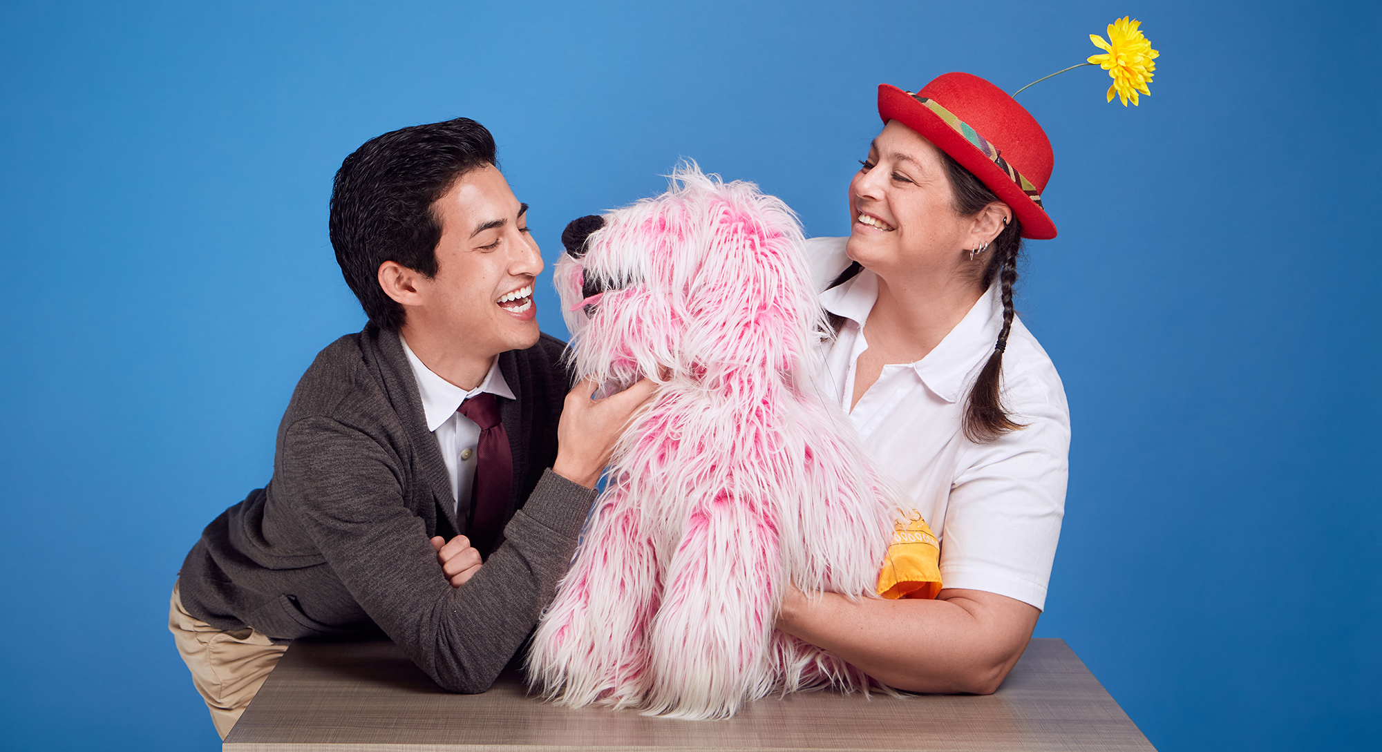 Photo of Flurffy the pink dog puppet with a smiling man on his left and a smiling woman on his right. The man wears a gray cardigan, white shirt, and dark red tie. The woman wears a white shirt and a red hat with a yellow flower on top.