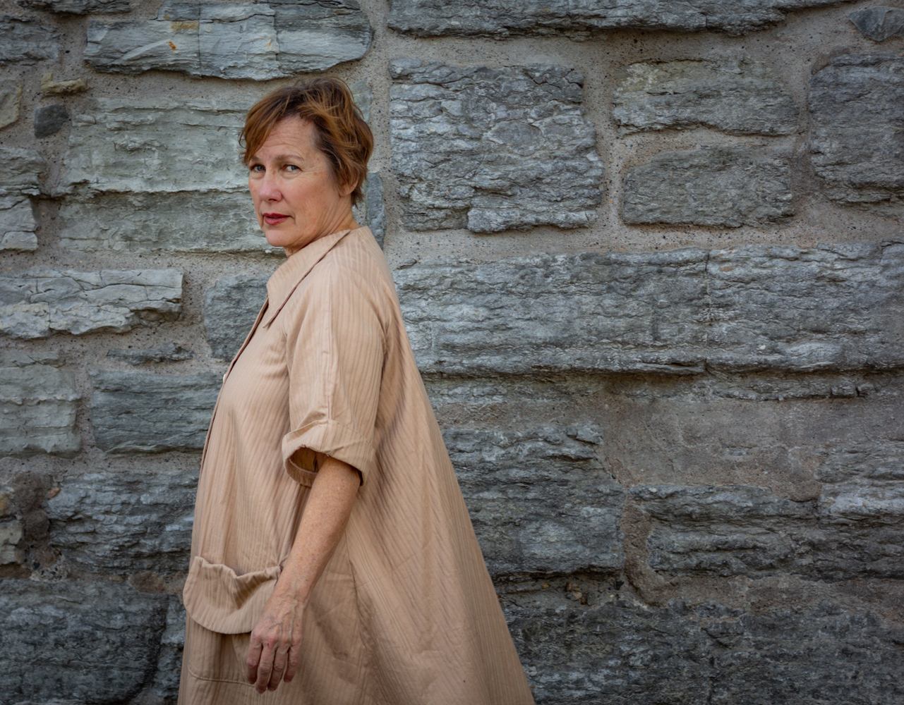 Photo of Iris Dement wearing a camel shirt dress walking to the left of the frame and looking at the camera sideways in front of a stone wall.