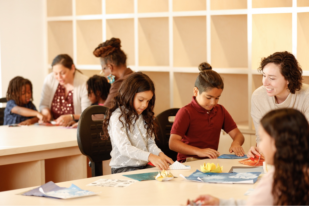 Photo of an art studio with two work surfaces. In the foreground, three kids and one teacher are sitting around one work station folding origami. In the background, there are two more students and two teachers around another work surface.