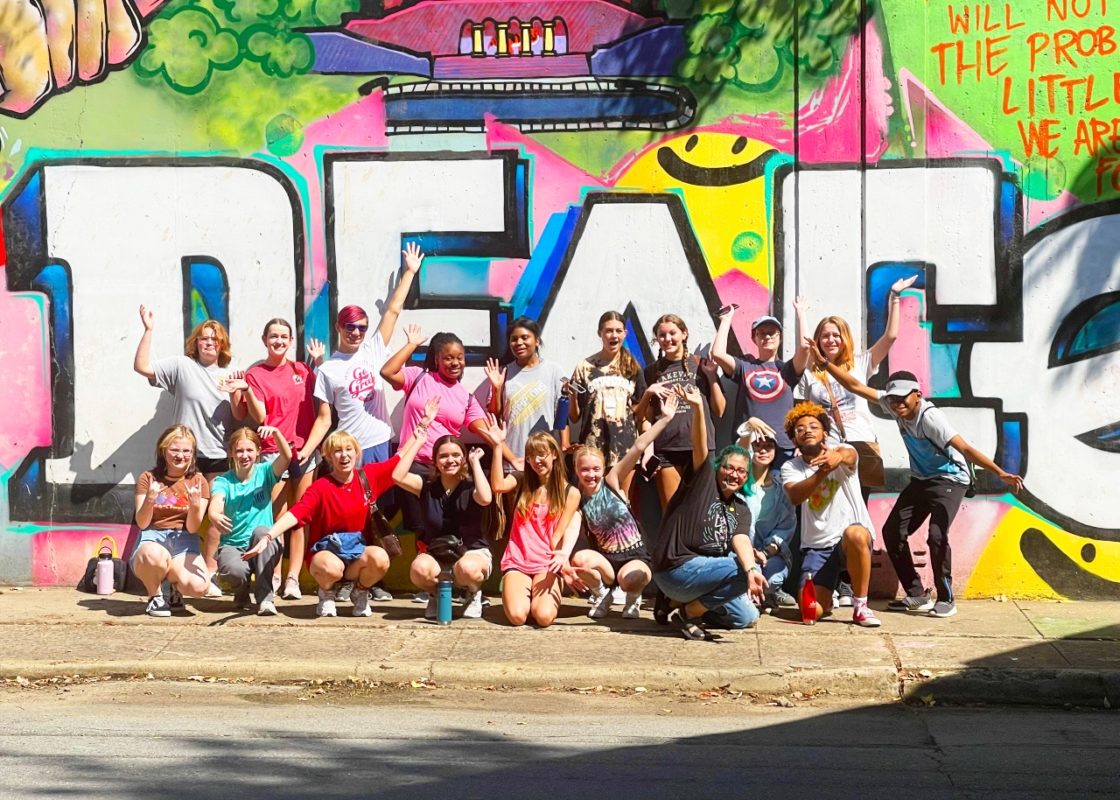 Photo of a group of teenagers posed in in front a wall mural making different faces and arm movements.
