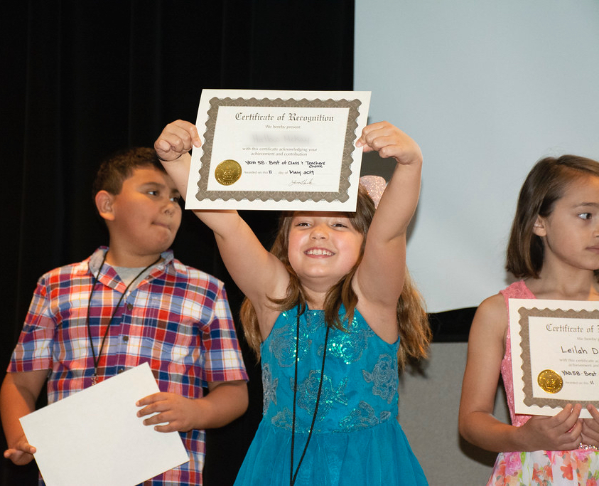 Photo of three small children. The girl in the middle is smiling and holding up a paper certificate reading "Certificate of Recognition."