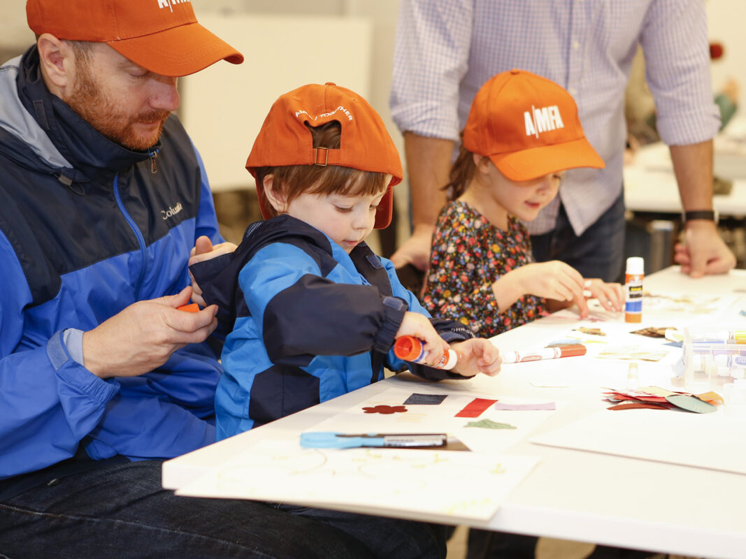 Photo of a man sitting with a young boy in his lap and a young girl next to him making arts and crafts at a white table. All three wear orange AMFA hats.