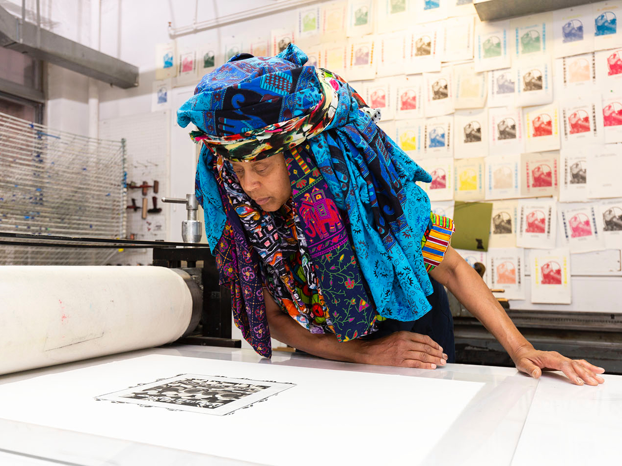 Photo of Chakaia Booker wearing a large blue headress in a workshop looking down at a print of her work on table in the foreground. There are dozens of color samples of a print on the wall behind her.