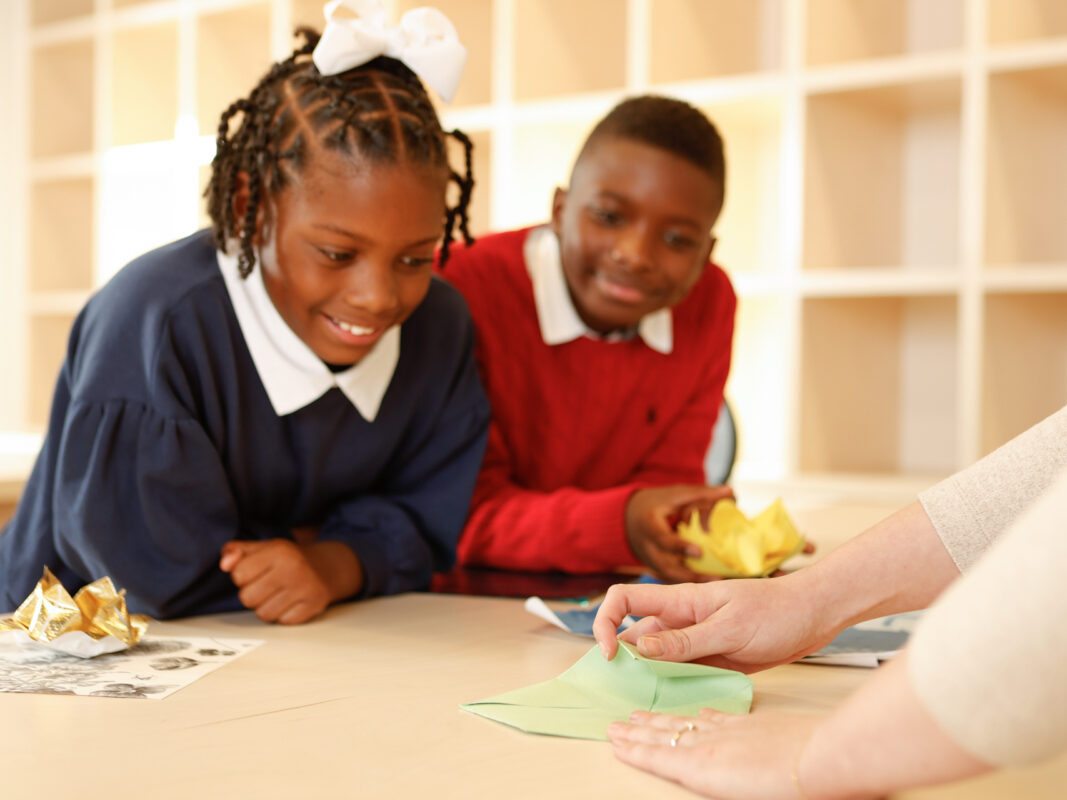 Photo of a girl and a boy in school uniforms leaning on a light wood table as a pair of adult hands demonstrates how to fold origami.