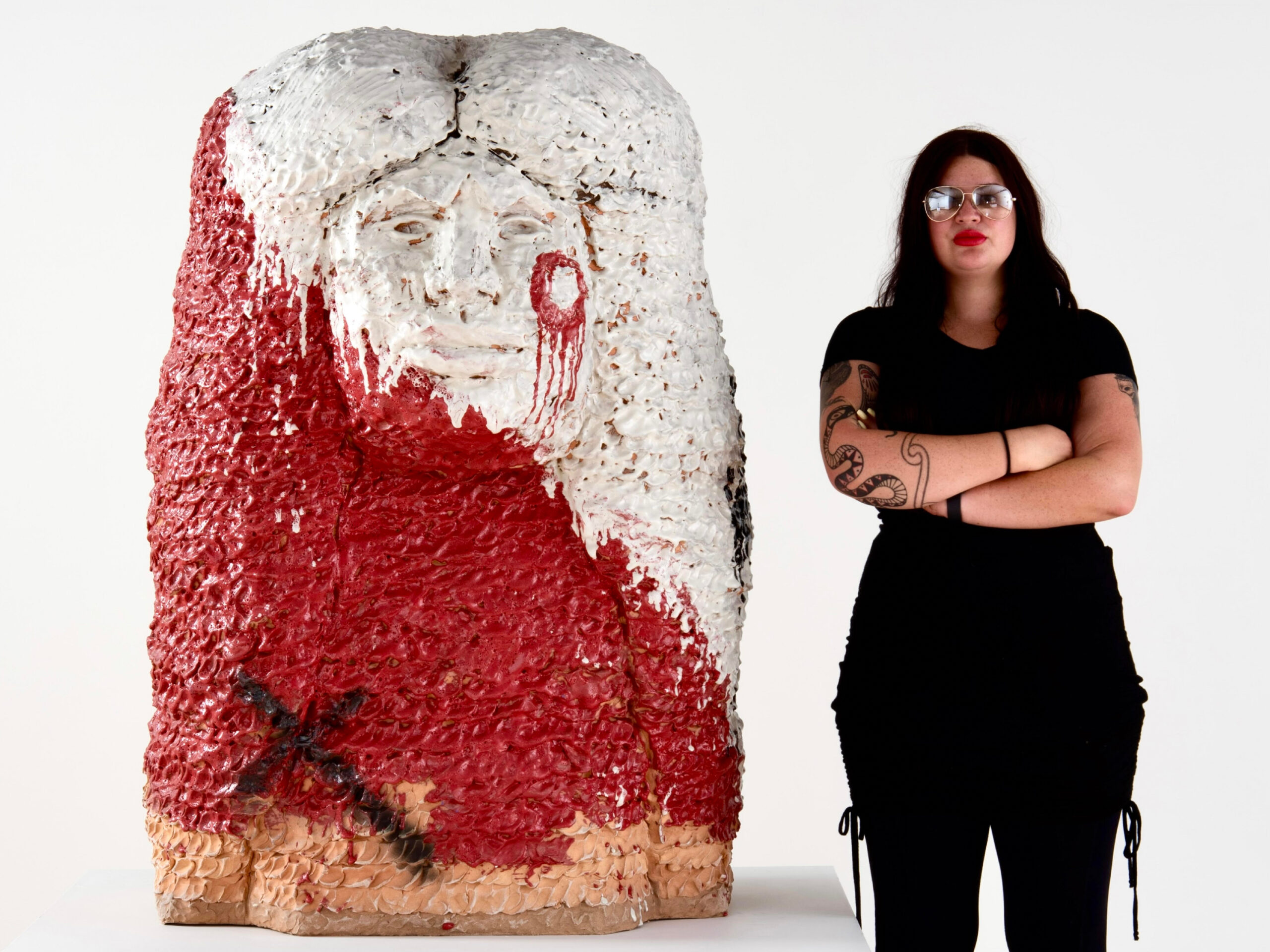 Photo of Raven Halfmoon wearing a black shirt and pants standing next to a large-scale ceramic sculpture of a head with long hair and blood-reed glaze and a black cross.