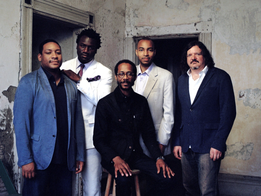 Photo of Brian Blade and the Fellowship Band - four men stand around one man sitting on a stool in an empty room with peeling plaster walls and two doorways in the background.
