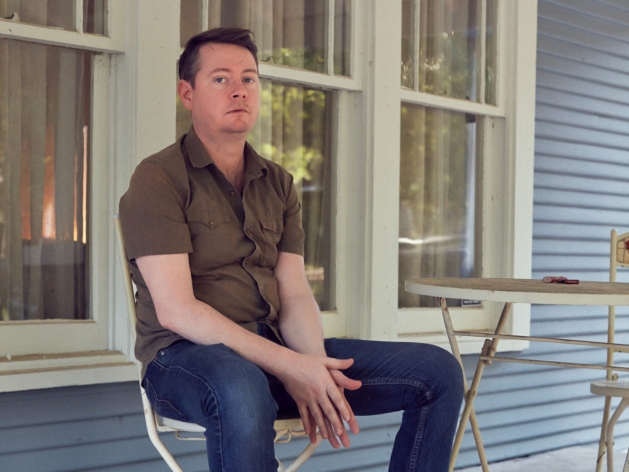 Photo of John Fullbright sitting in a white metal chair in front of a three windows with white frames on a house with blue siding.