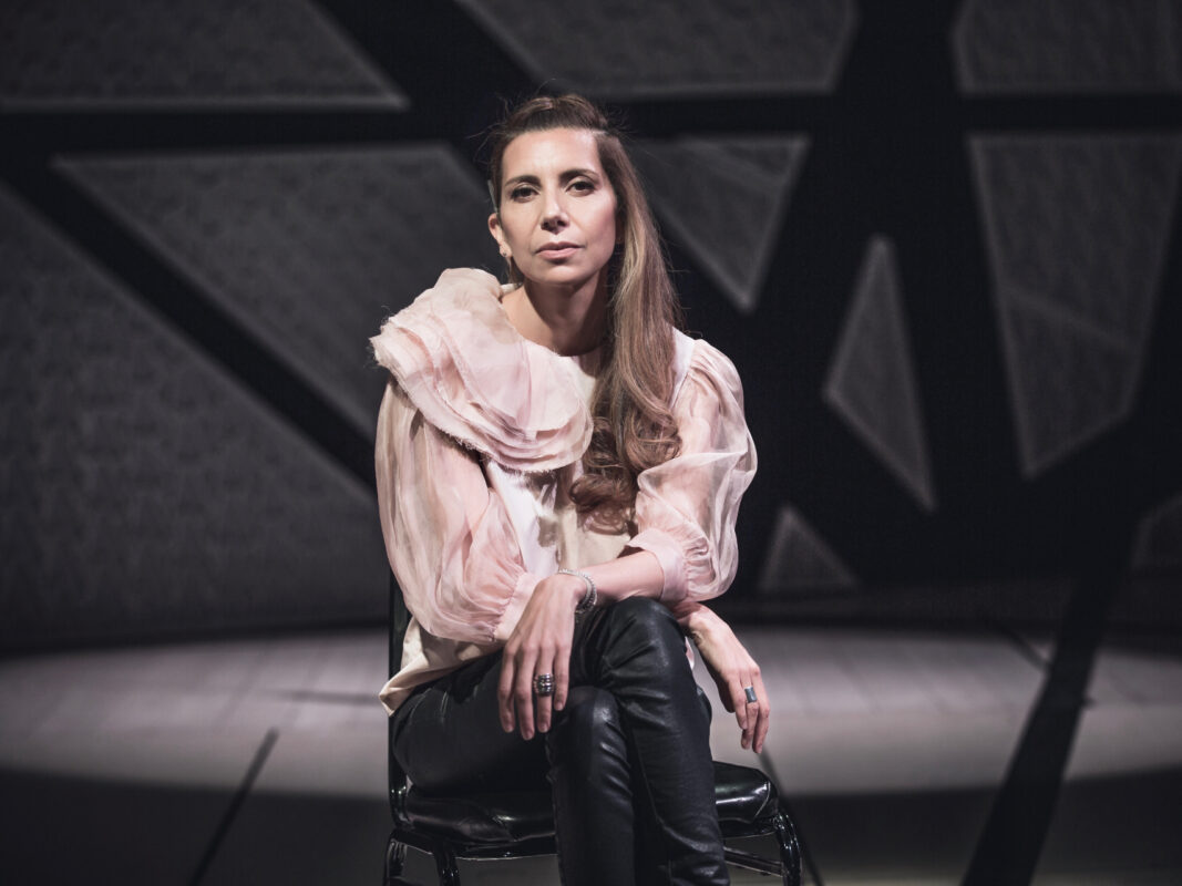 Photo of Magos Herrera wearing an pink blouse and black leather pants sitting on a black stool in front of an empty stage.