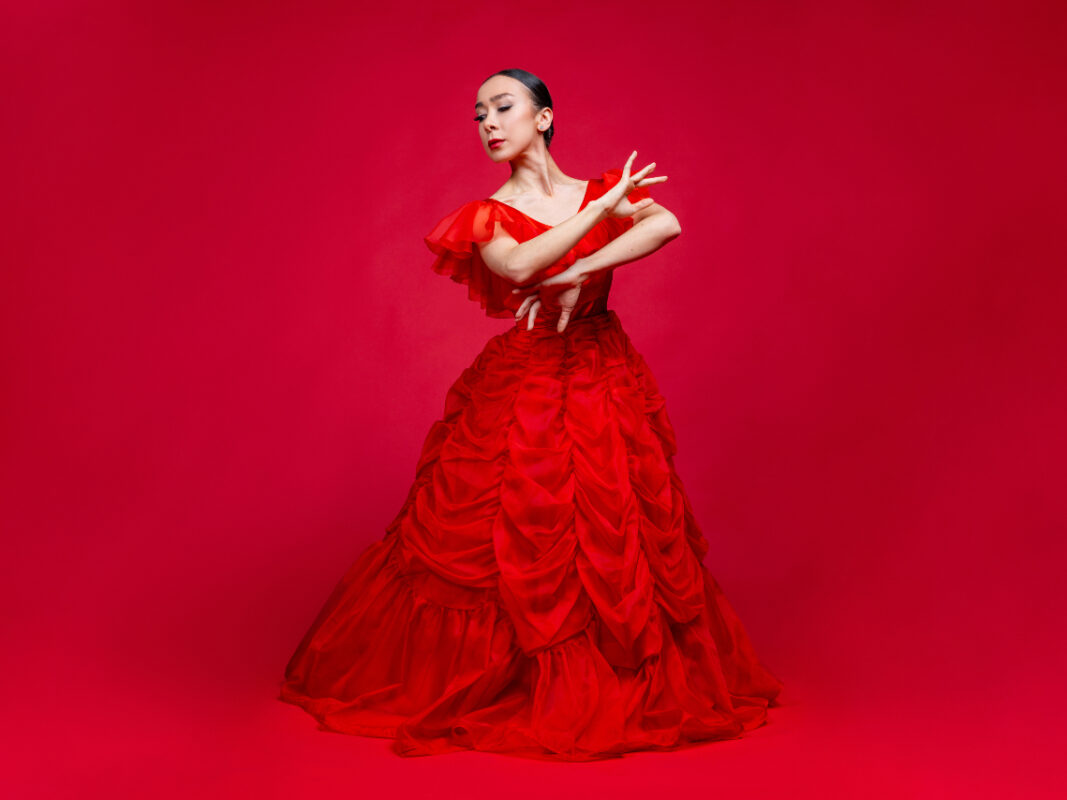 Photo of a female dancer wearing a red ballgown while posed in front of a red background.