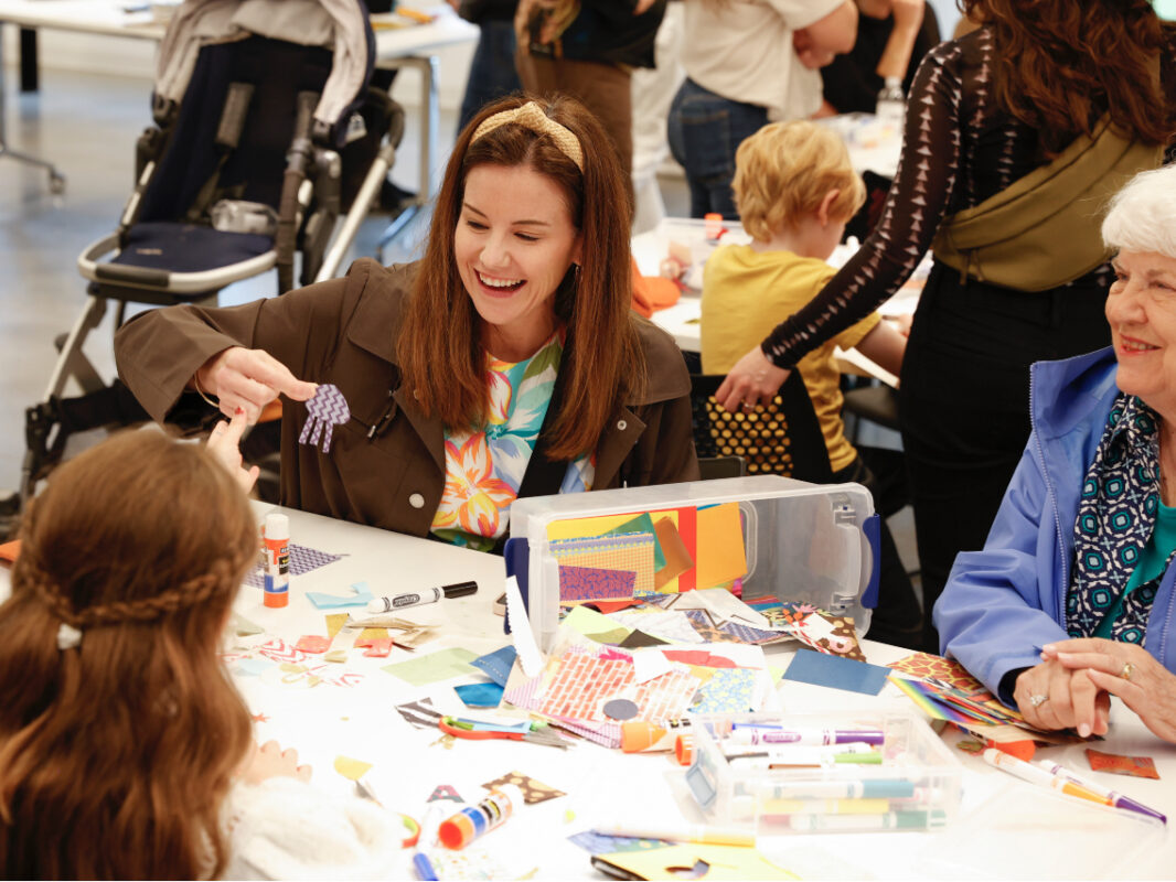 Photo of a mother, grandmother, and young girl sitting at a table doing arts and crafts with other families in the background.