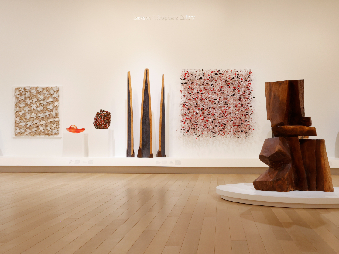 Photo of a large modern wooden sculpture in front of a wall with other sculptures in an art gallery.