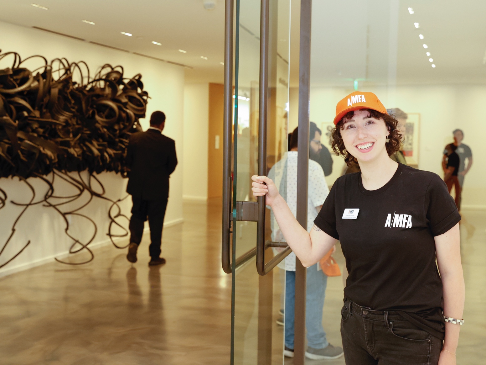 Photo of a woman wearing an AMFA T-shirt and hat holding open the door to an art gallery.
