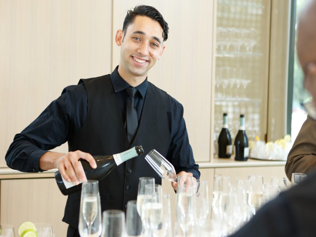 Photo of a bartender wearing a black shirt and tie pouring a glass of champagne.