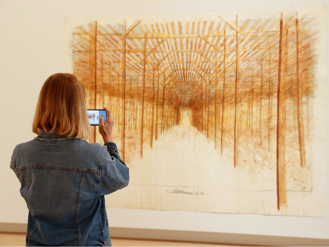 Photo of a woman viewed from behind standing in front of a painting while holding up her cell phone to take a picture.