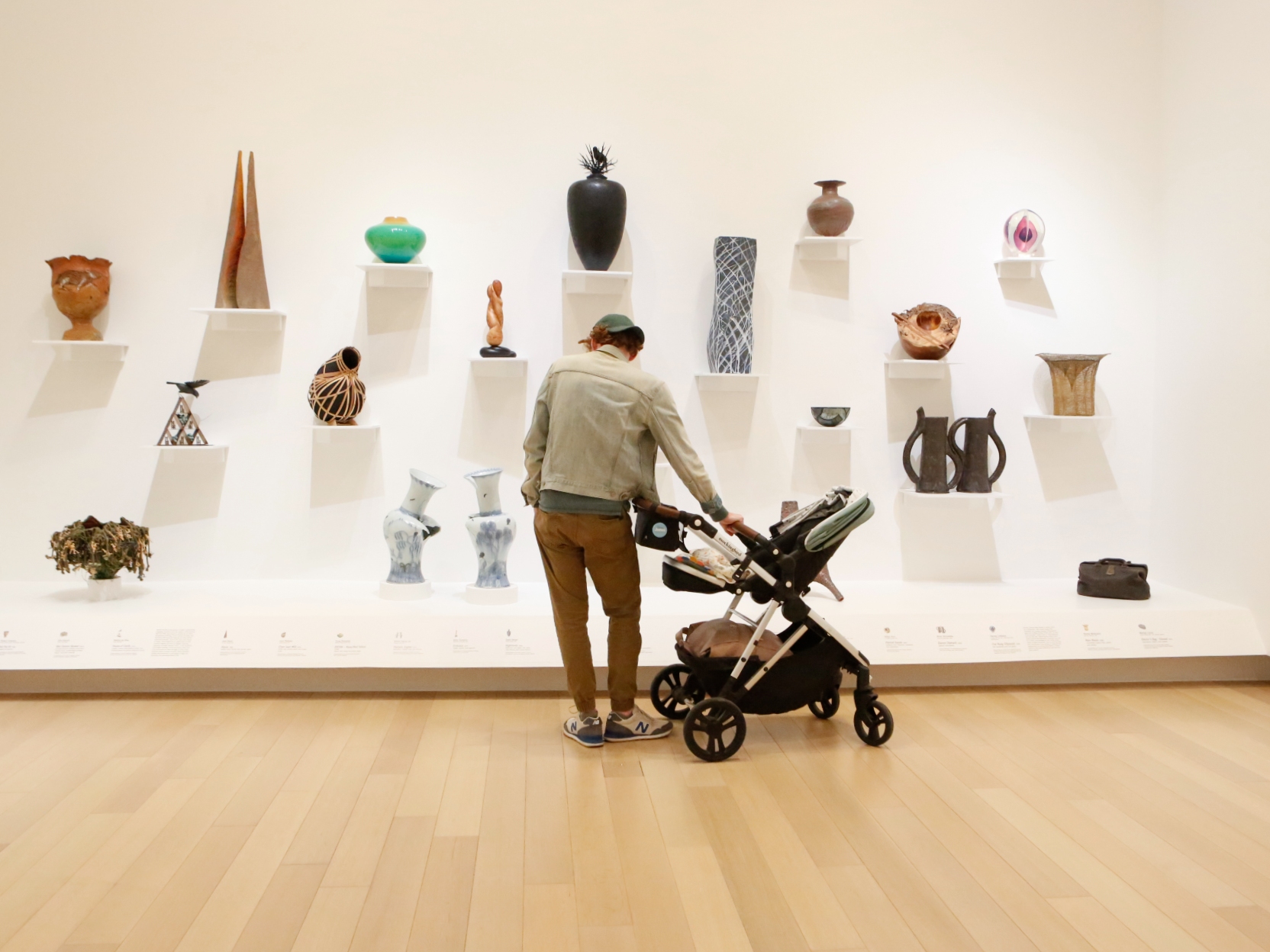 Photo of a man pushing a stroller in front of a wall in an art gallery displaying small ceramics on individual shelves.
