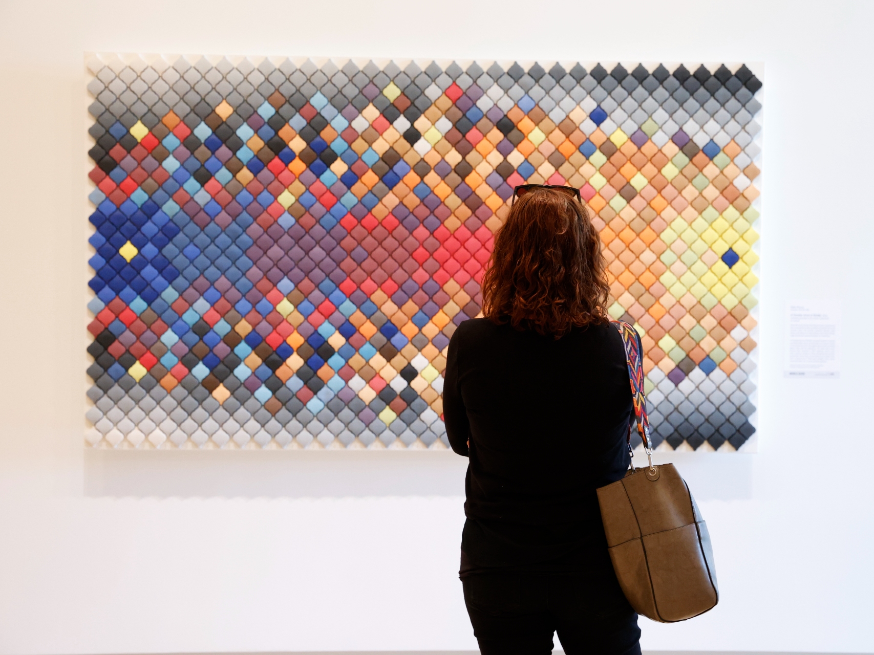 Photo of a woman with her back to the camera observing a brightly-colored tile artwork on the wall of an art gallery.