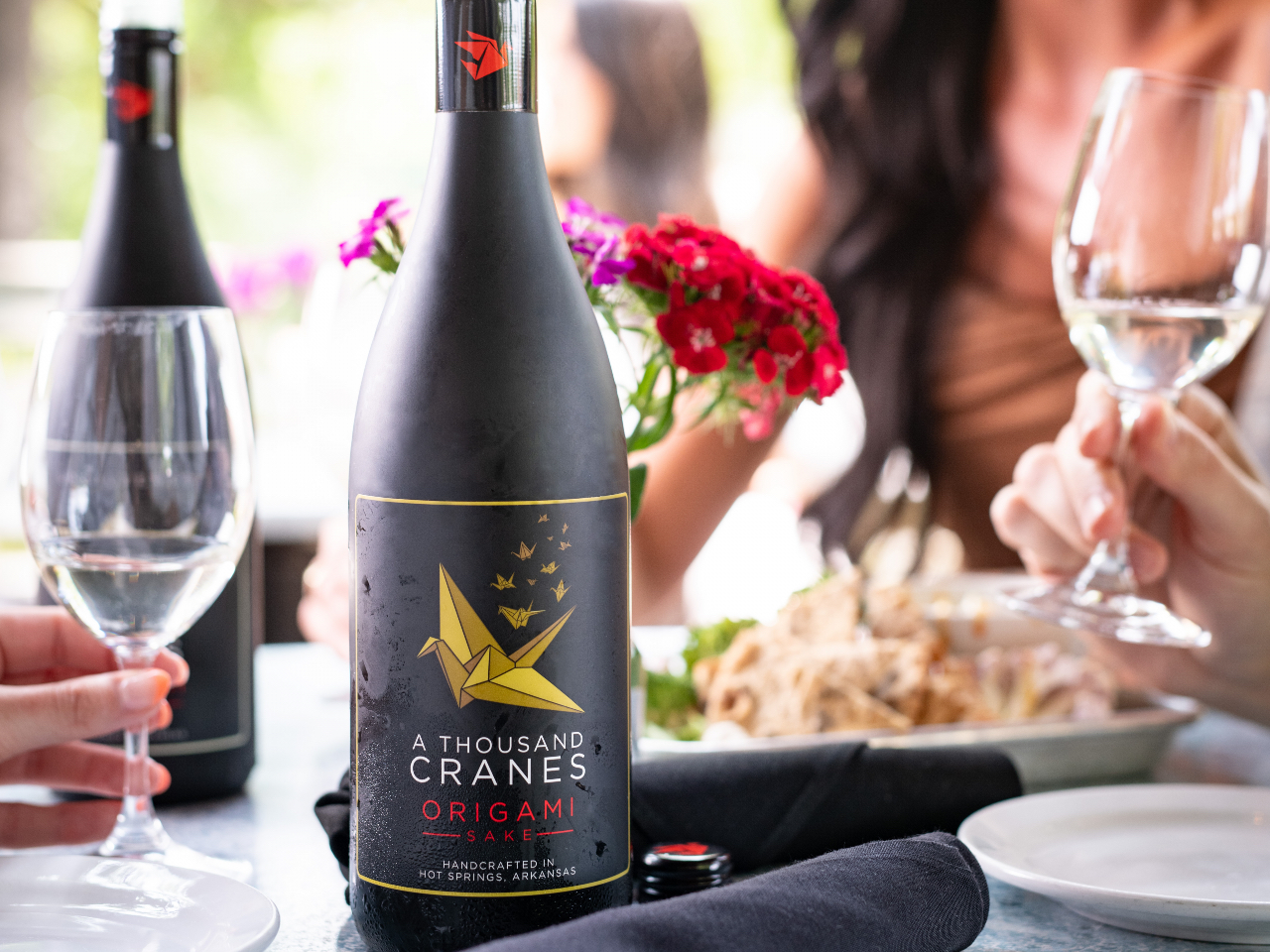 Close-up photo of a bottle of A Thousand Cranes Origami Sake on a table with people holding glasses in the background.