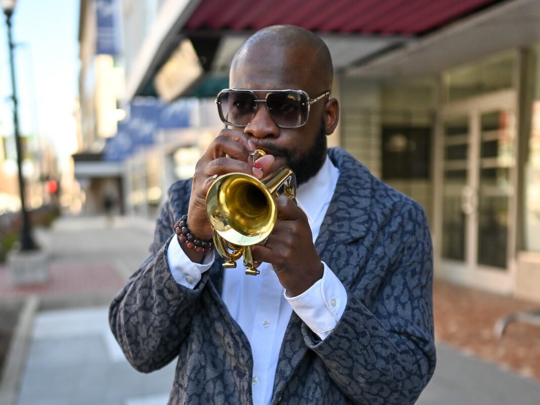 Photo of Rodney Block playing a trumpet on a sidewalk next to a buliding.