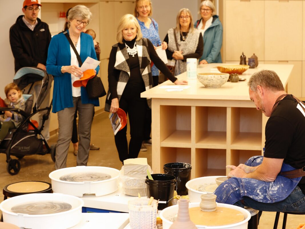 Photo of a man throwing pottery on a wheel in an art studio while a group of people watch.