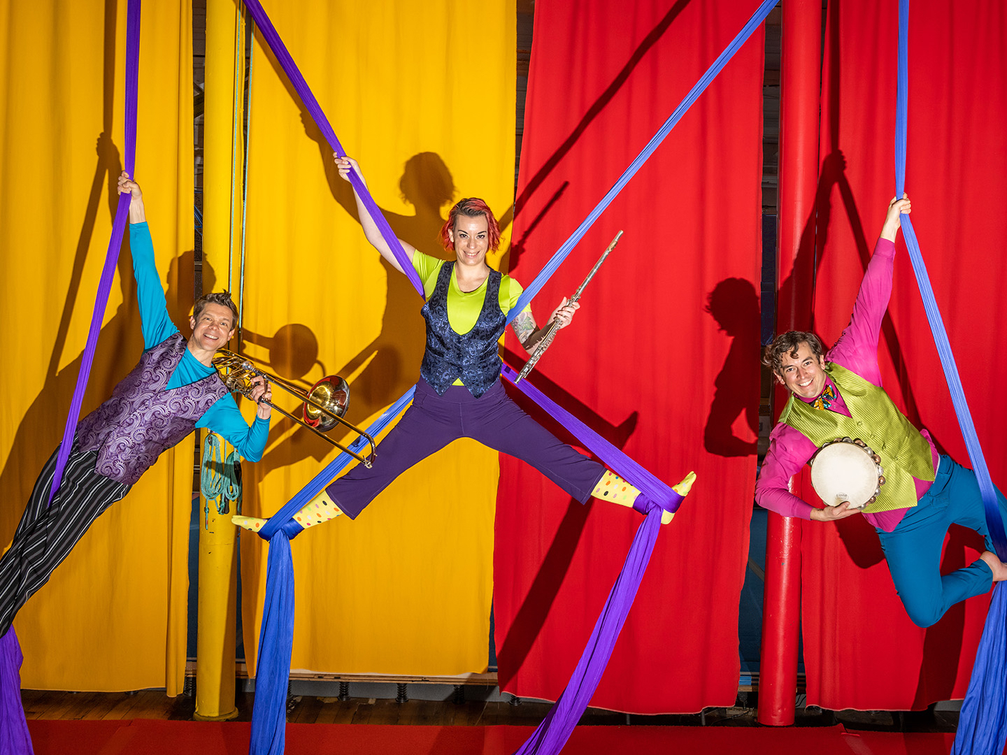 Photo of a trumpet player, flute player, and tambourine player hanging from purple silks above a stage with red and yellow curtains behind them.