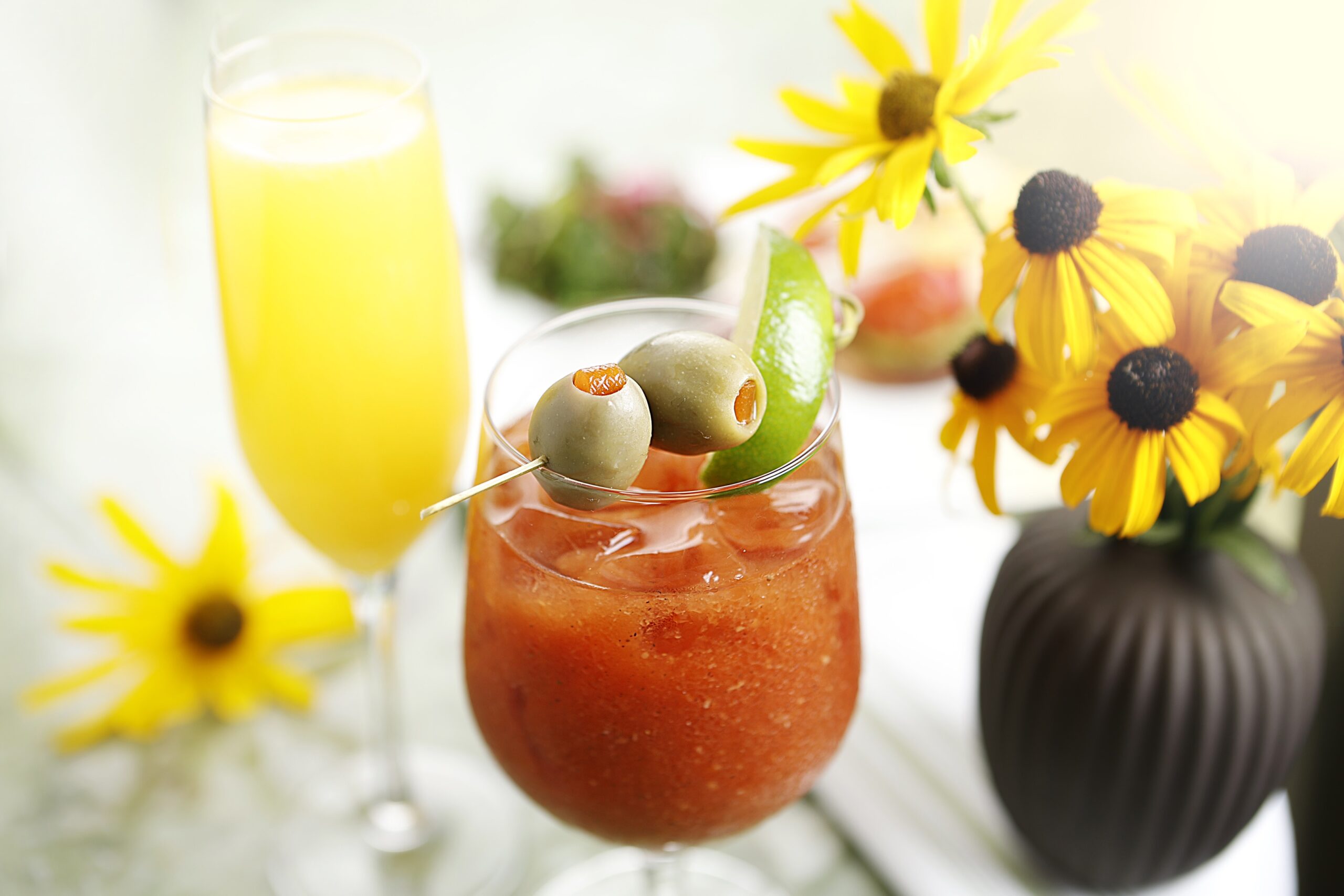 Photo of a Bloody Mary and Mimosa on a taptop next to a vase of yellow flowers.