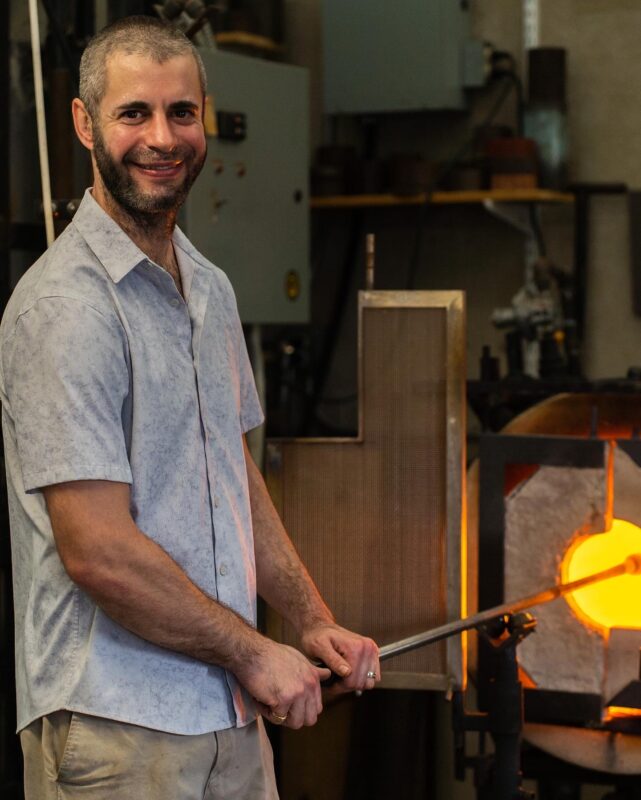 Photo of Ben Dombey holding a rod with molten glass on the end over a fire.