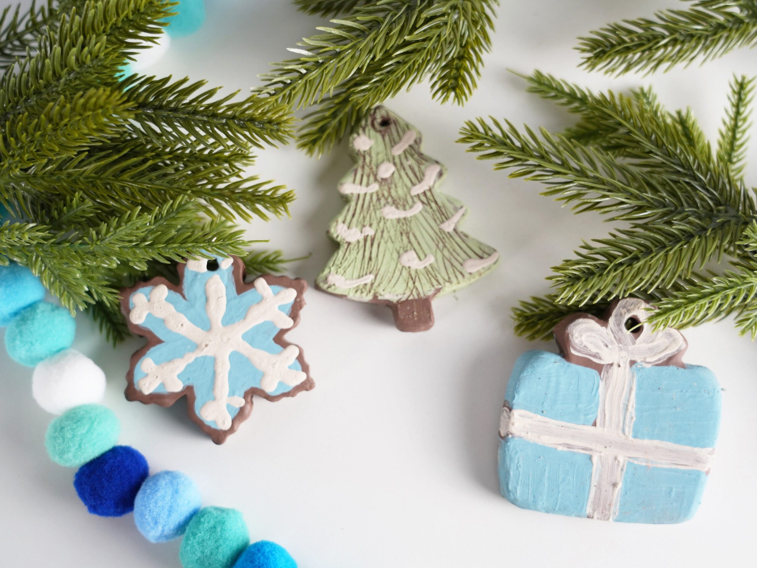 Photo of clay ornaments shaped like a snowflake, Christmas tree, and wrapped gift hanging on tree limbs.