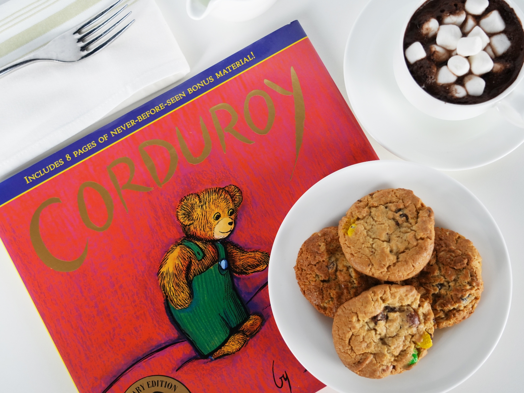 Photo of the book 'Corduroy,' a plate of cookies, and a cup of hot chocolate on a white tabletop.