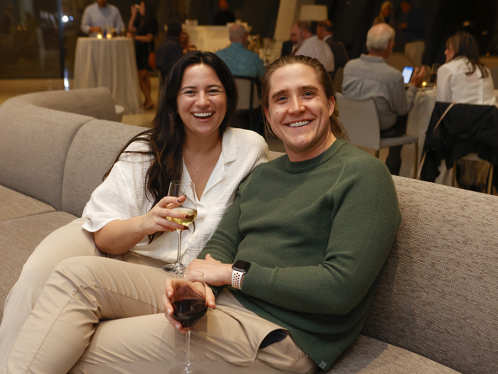 Photo of a man and woman smiling and holding wine glasses while seated on a gray couch in the Cultural Living Room.