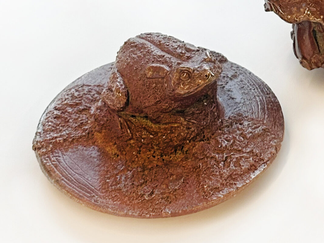 Close up photo of a glazed brown clay pot lid with a small sculpture of a frog on the surface.