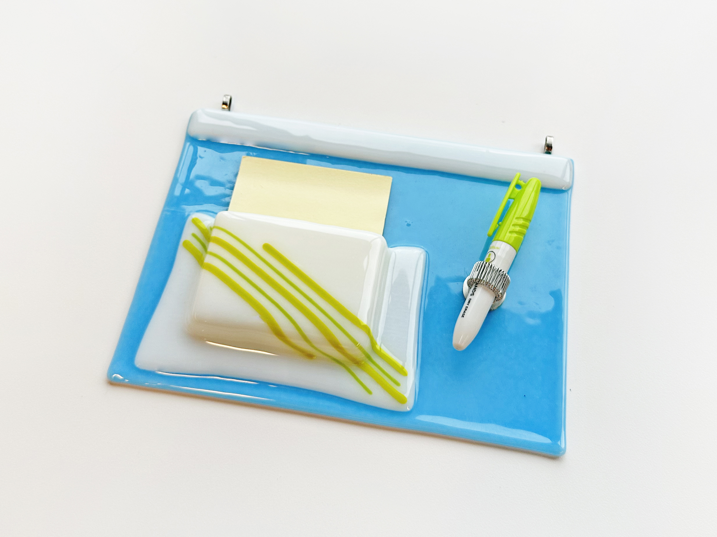 Photo of a fused glass wall hanging that resembles a tabletop with a folder for post it notes and a silver loop for a green sharpie pen.