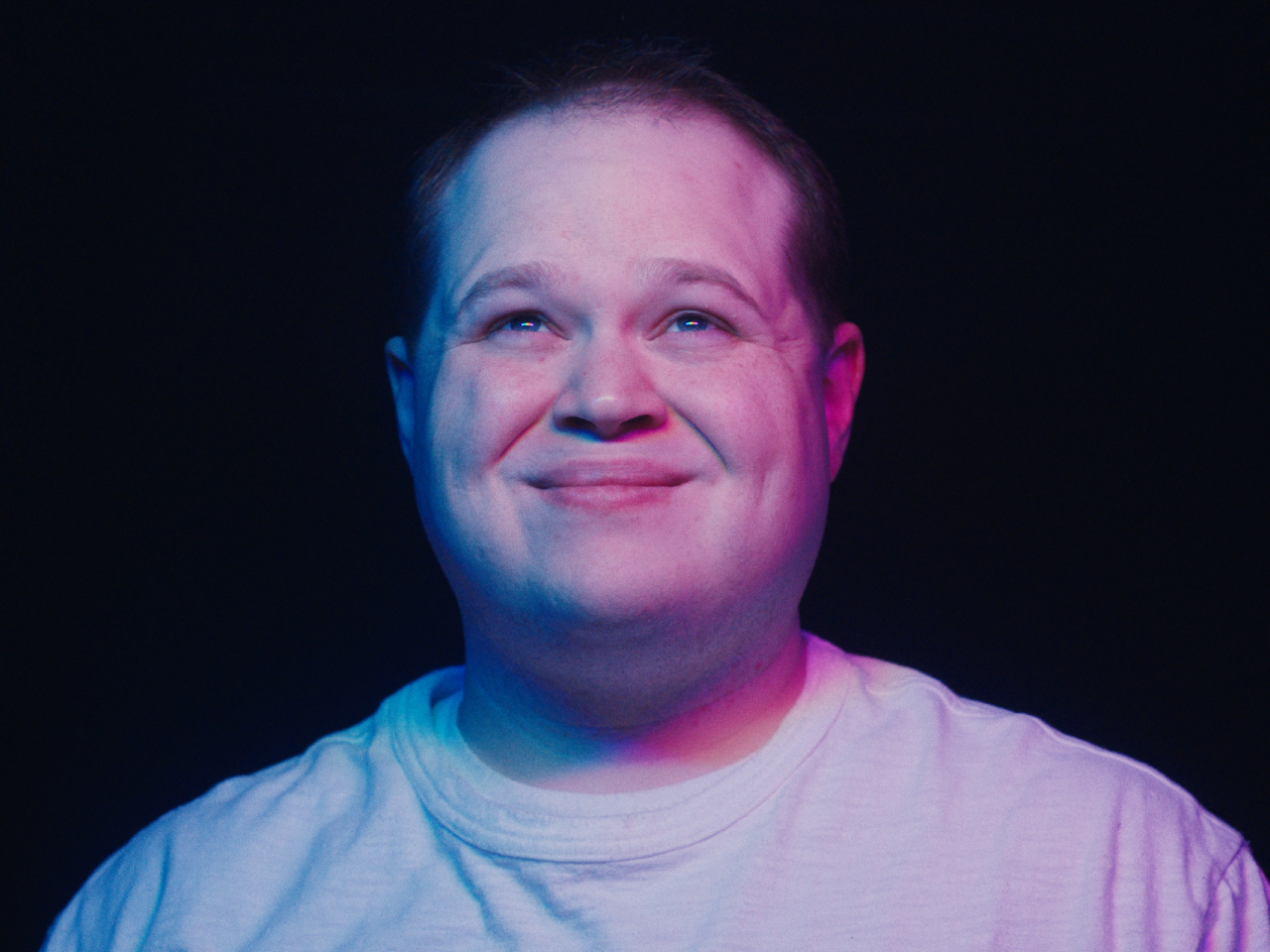 Film still from You Have No Idea depicting a man smiling with a blue and purple light shining on his face.