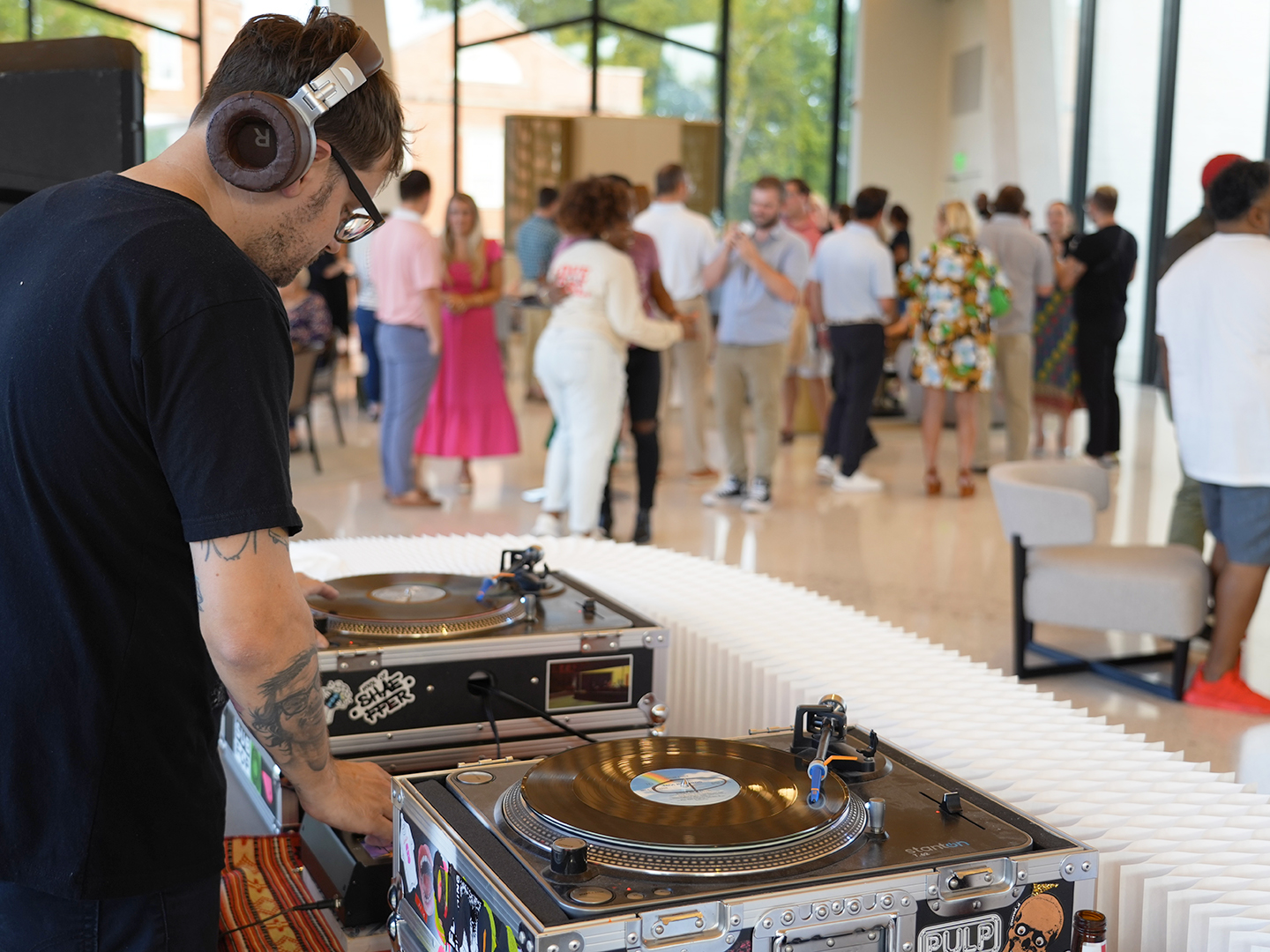 Photo of a DJ mixing records on a set of turntables with groups of people mingling and dancing at a party in the background.