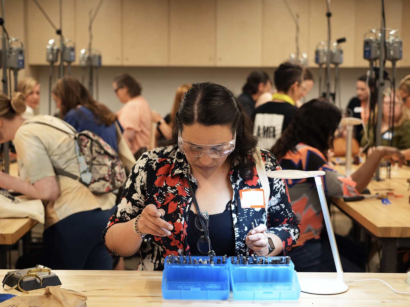 Photo of a woman wearing goggles and looking in a blue box of metal tools in an art studio where other people are interacting in the background.