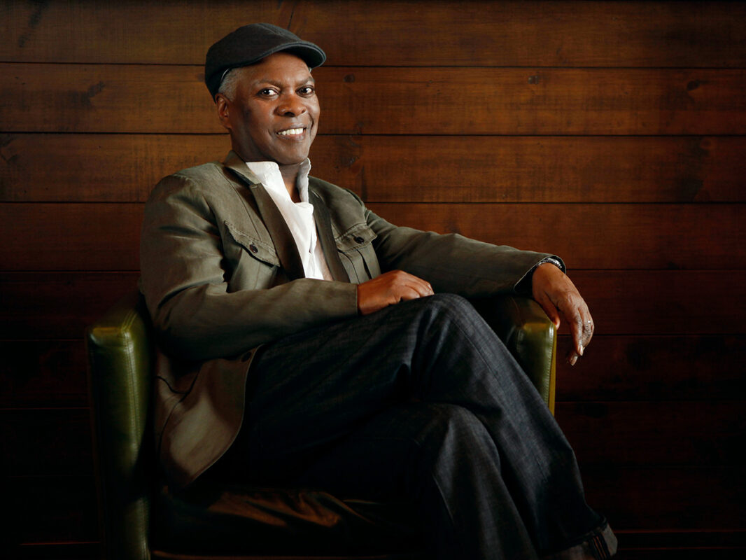 Photo of Booker T wearing a green jacket white shirt and dark jeans sitting on a green chair in front of a wood paneled wall.