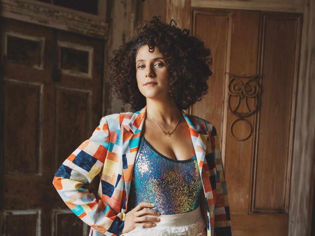 Photo of a woman with brown curly hair wearing a multi-colored jacket, sequin top, and white shorts posed with her hand on her hip.