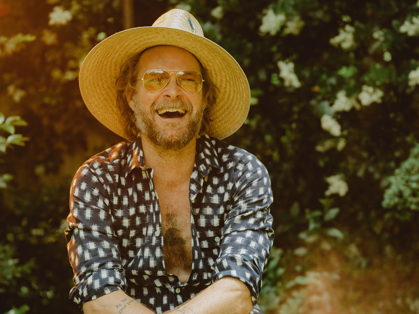 Photo of a man smiling while wearing a straw hat, yellow sunglasses, and a open blue button down shirt in front of a wooded area.