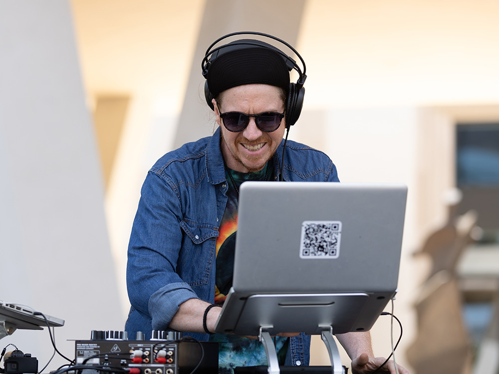 Photo of DJ Mike Poe wearing headphones and sunglasses while typing on a laptop behind a DJ booth.