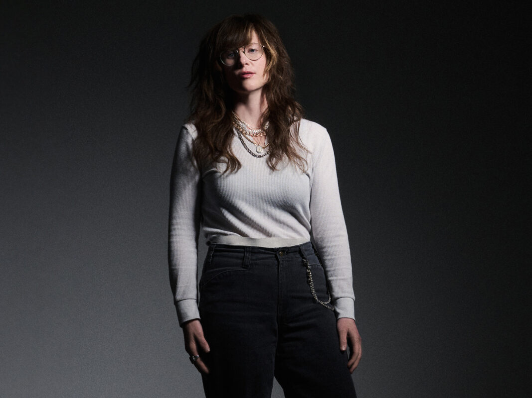 Photo of a woman wearing glasses, several beaded necklaces, a white shirt, and black pants standing in front of a gray background.