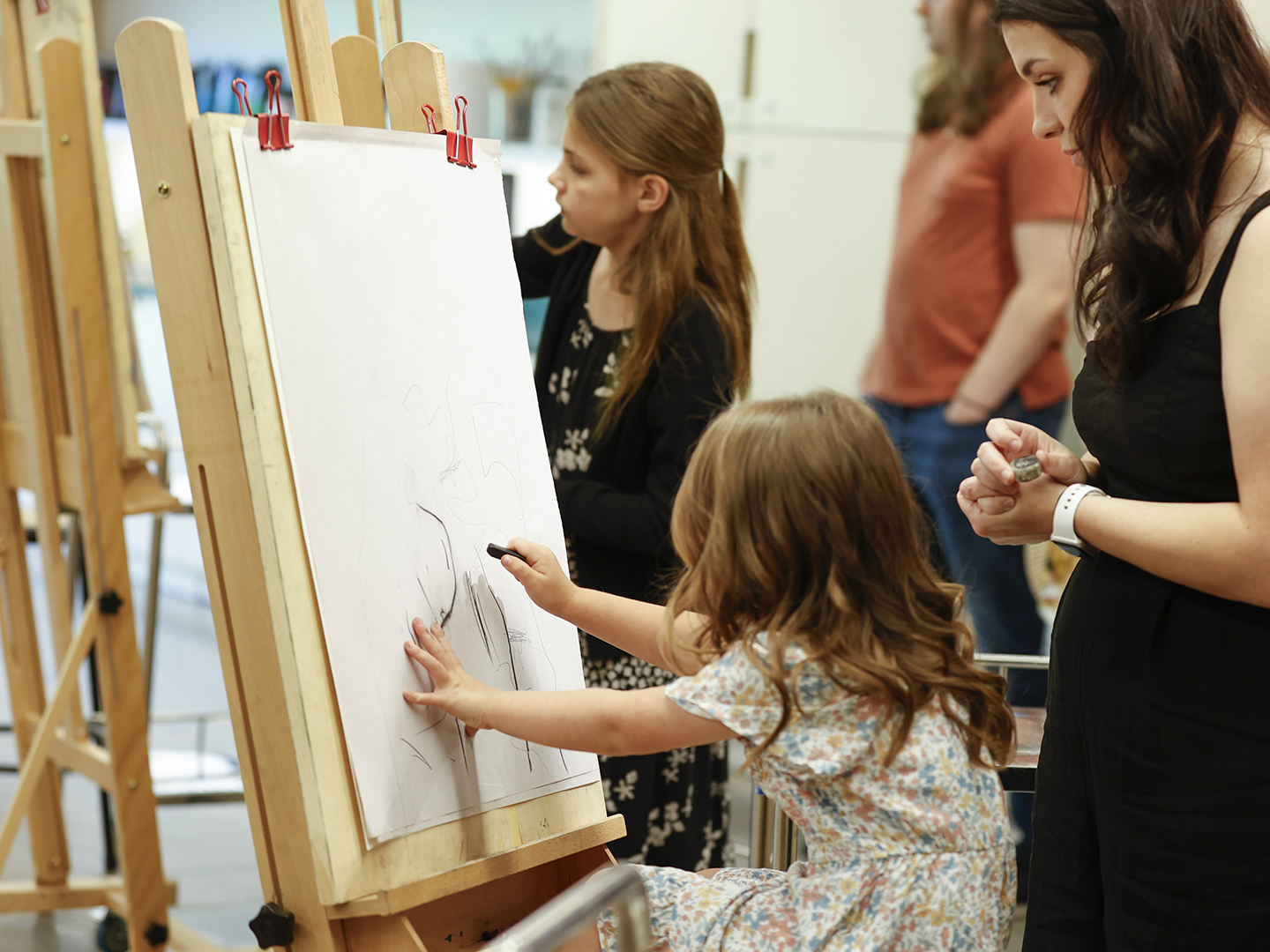 Photo of two young girls drawing at easels while adults look over their shoulders.