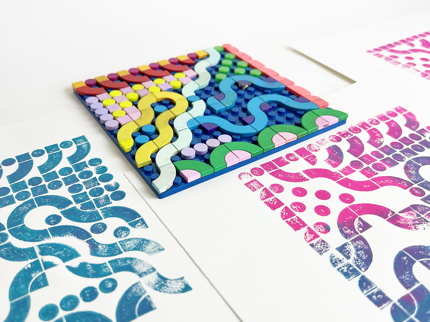 Photo of a few colorful handmade prints and a lego block stencil laying on a white surface.