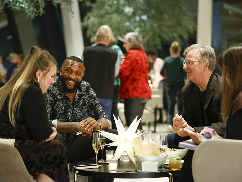 Photo of two men and two women sitting an a table drinking champagne with other people in the background.