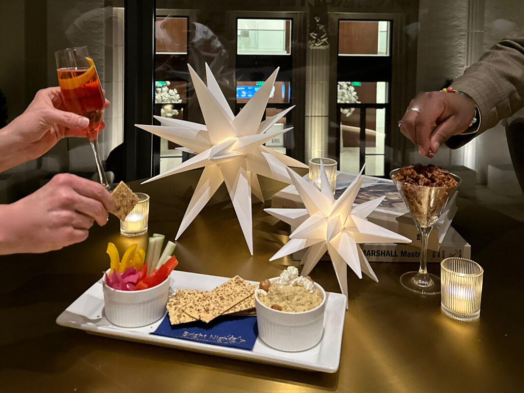 Photo of two sets of hands holding cocktails and reaching for snacks on a table decorated with votive candles and paper stars.