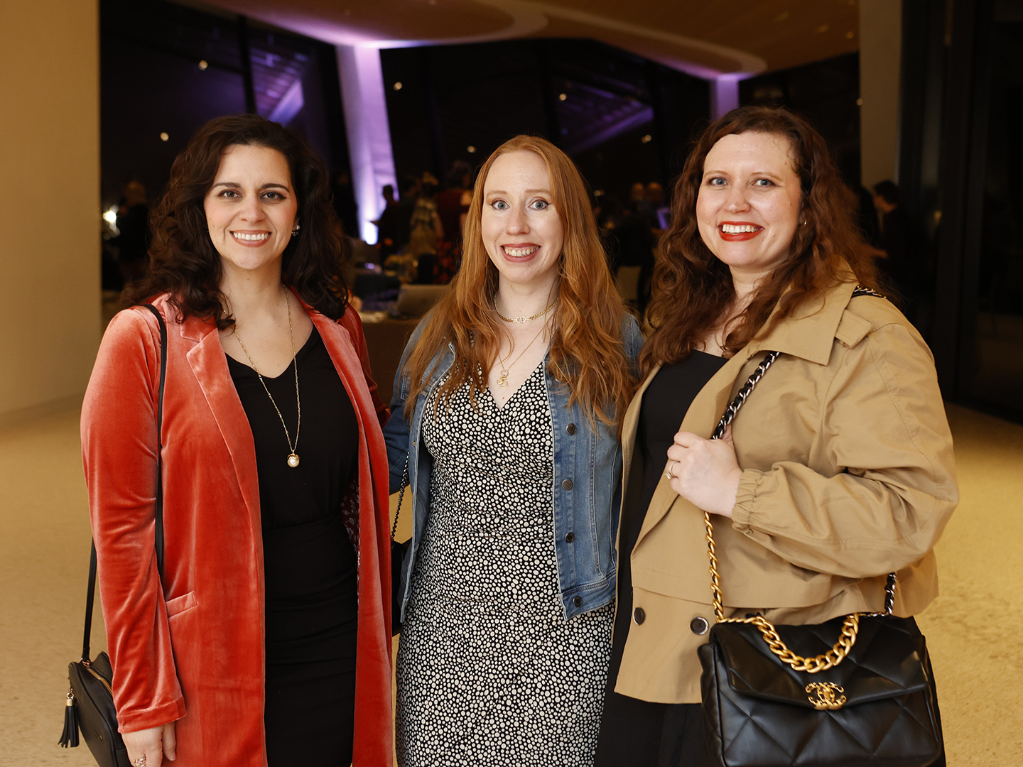 Photo of three women posed and smiling with a crowd of people at a dark lit party behind them.