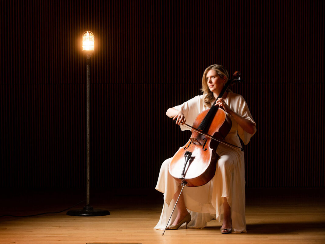 Photo of a seated woman wearing a white dress and playing a cello on a dark stage illuminated by a single light bulb.