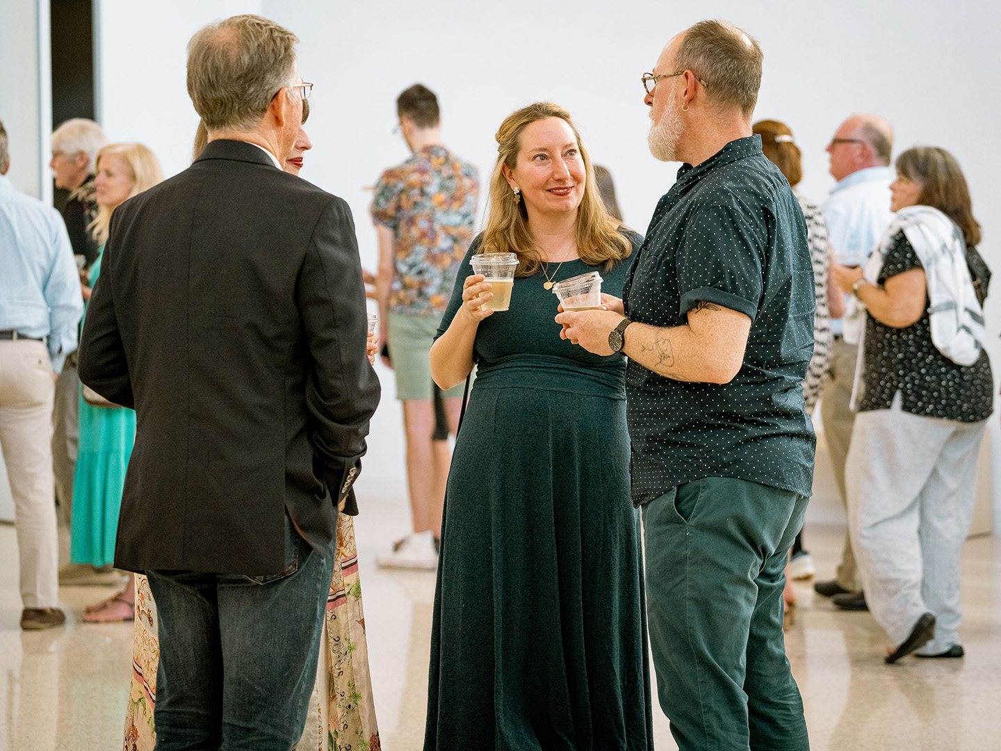 Photo of two women and two men standing in the Atrium of AMFA, holding plastic cups of wine, and talking to each other with a crowd of other people in the background.