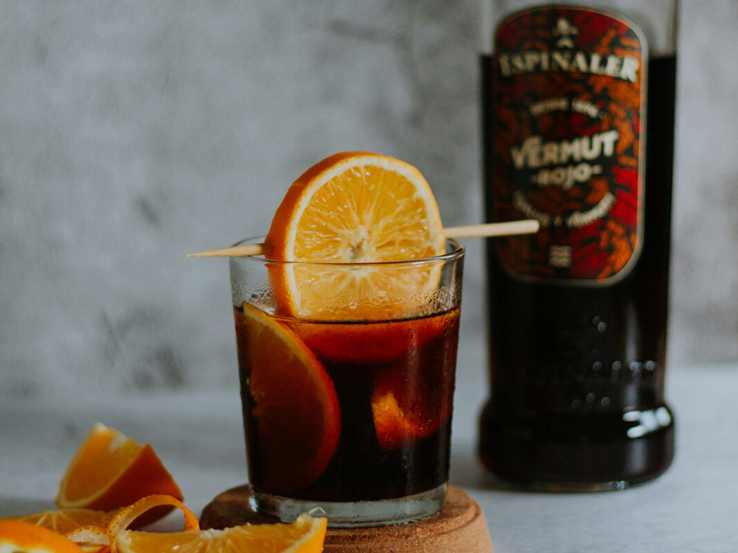 Photo of a dark brown cocktail garnised with orange in a rocks glass sitting next to a bottle of vermouth.